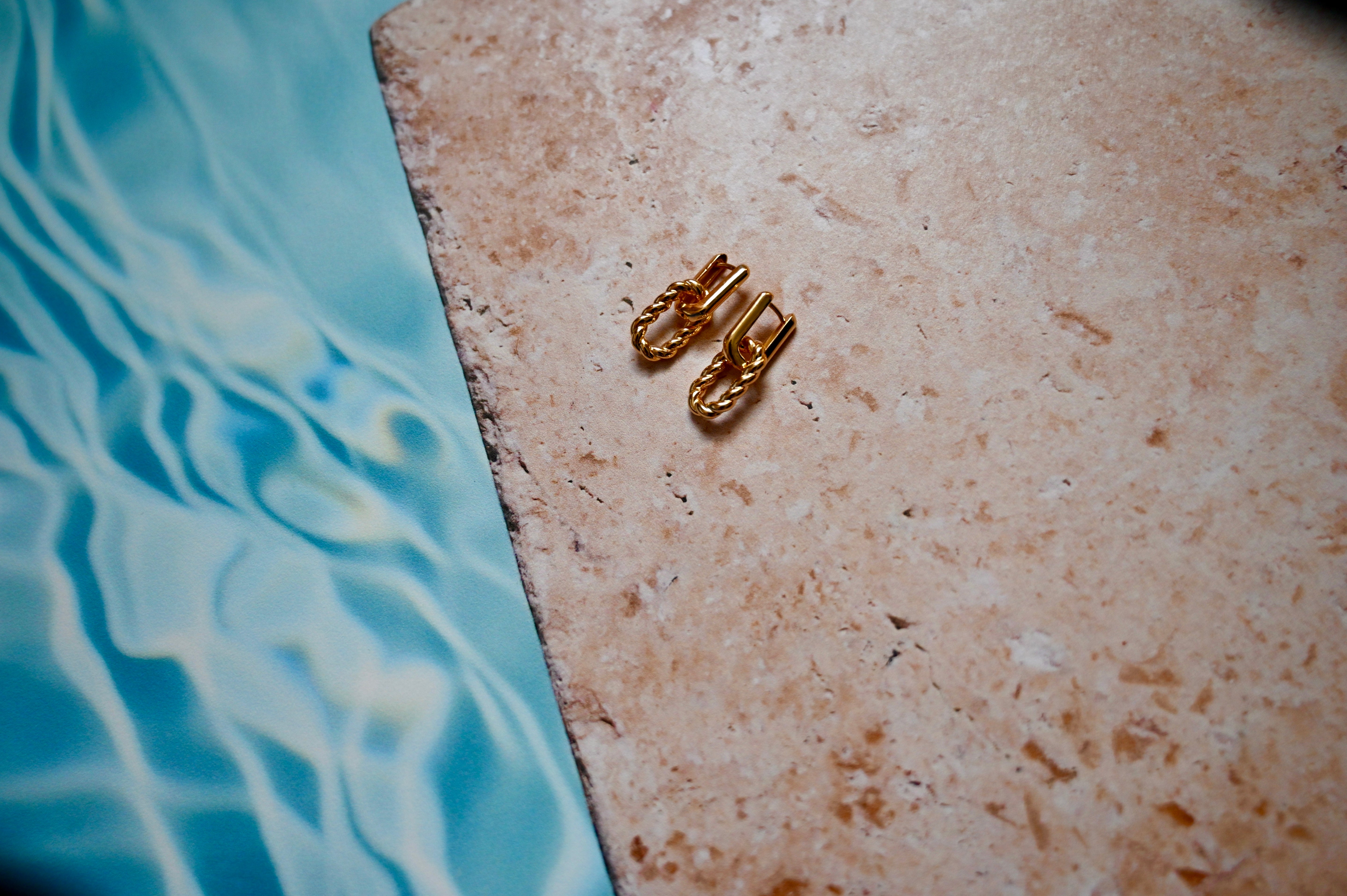Two gold chain link earrings on a textured surface with water in the background