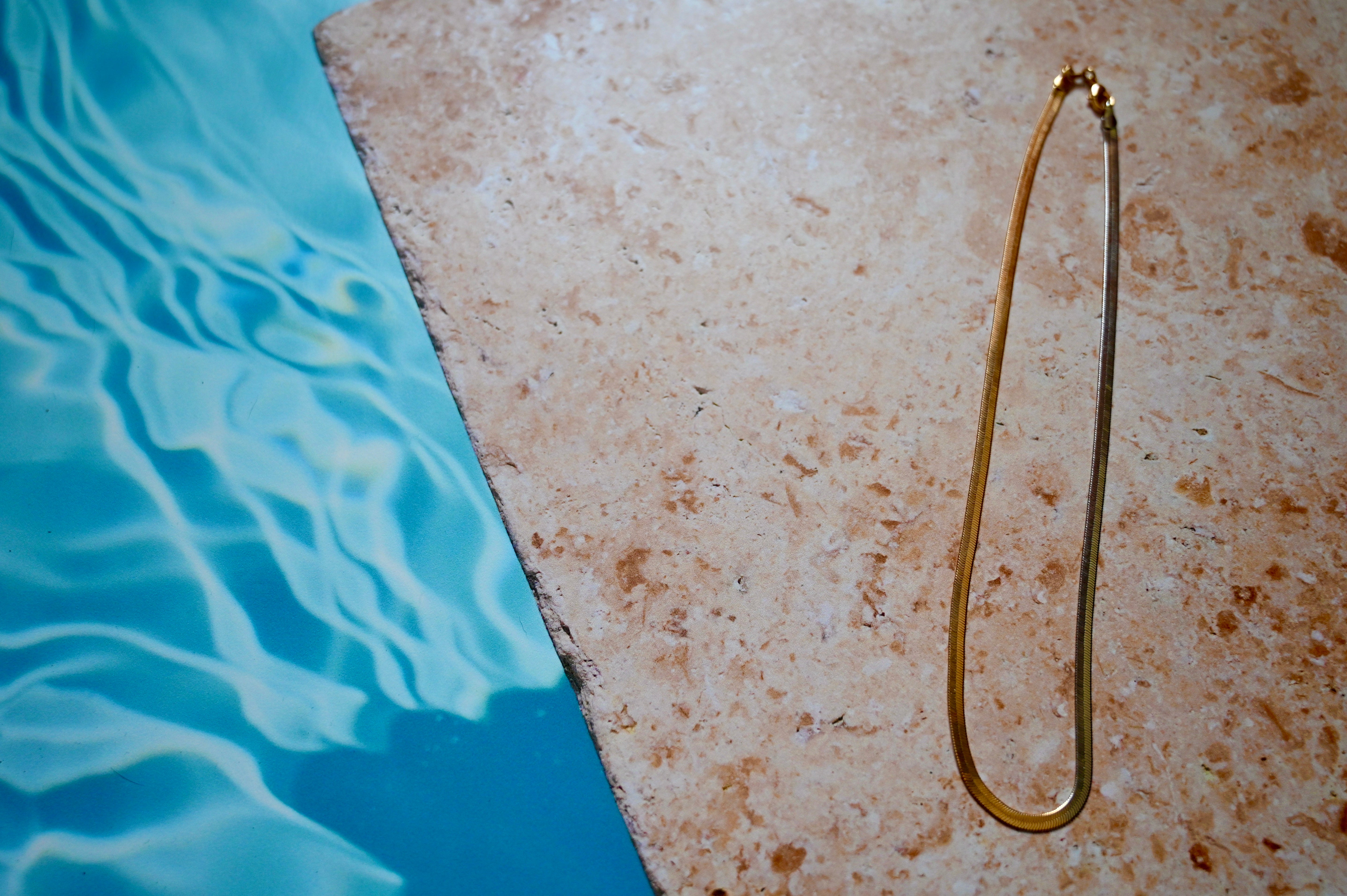 Gold and silver herringbone chain necklace laying on a stone by a pool.
