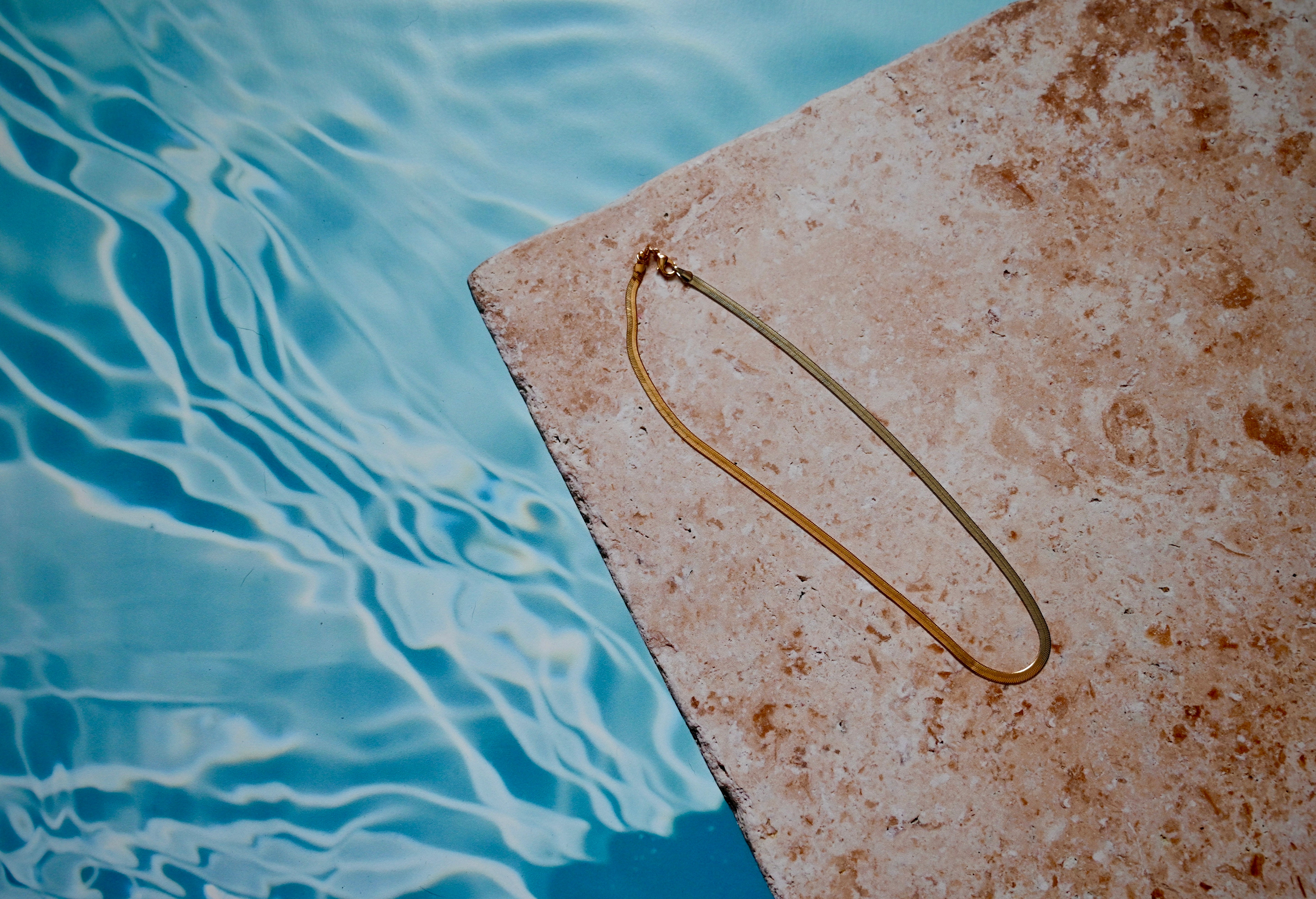 Gold and silver herringbone chain necklace laying on a stone by a pool.