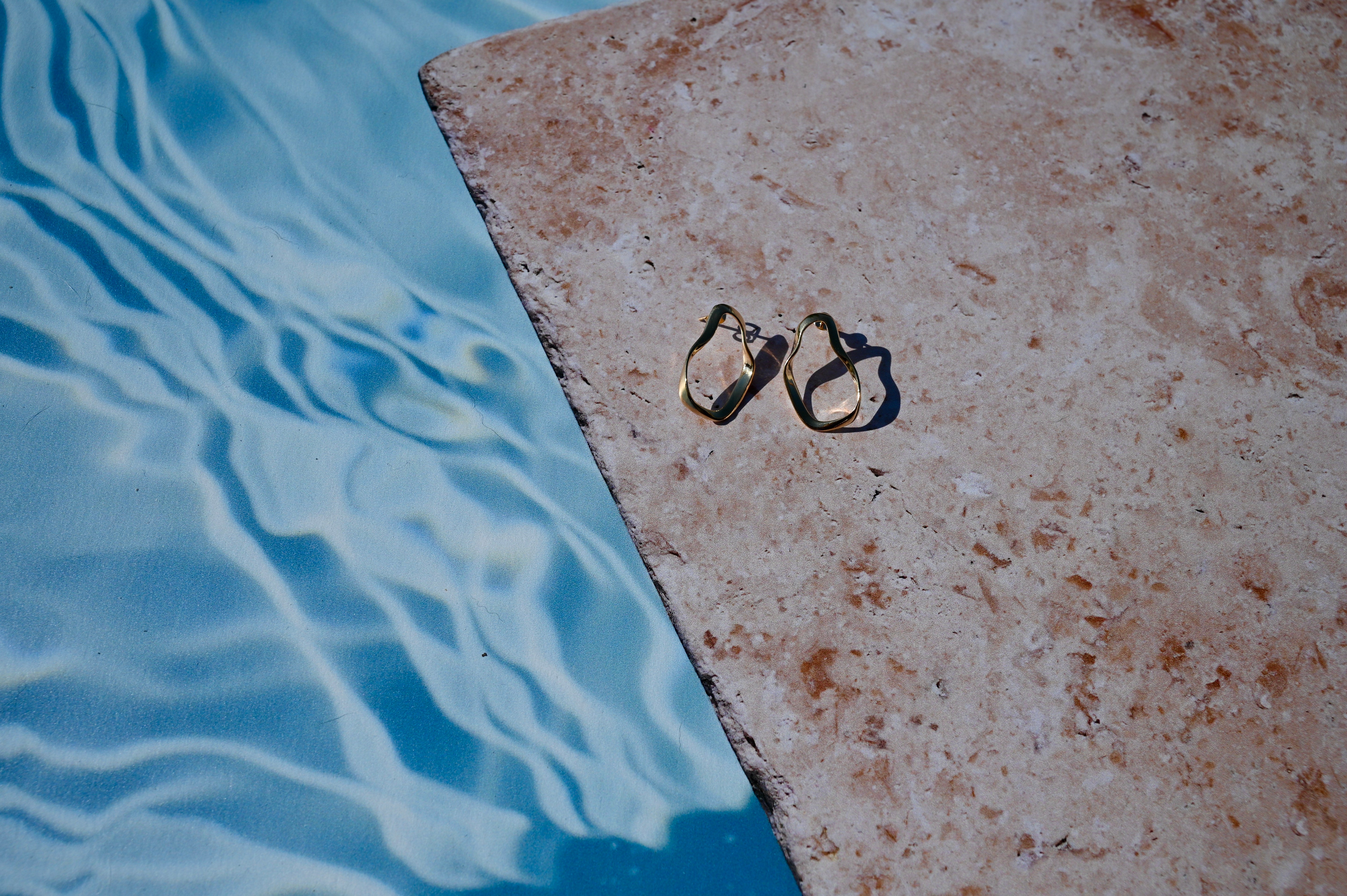 Asymmetric gold hoop earrings on a stone ledge by a pool