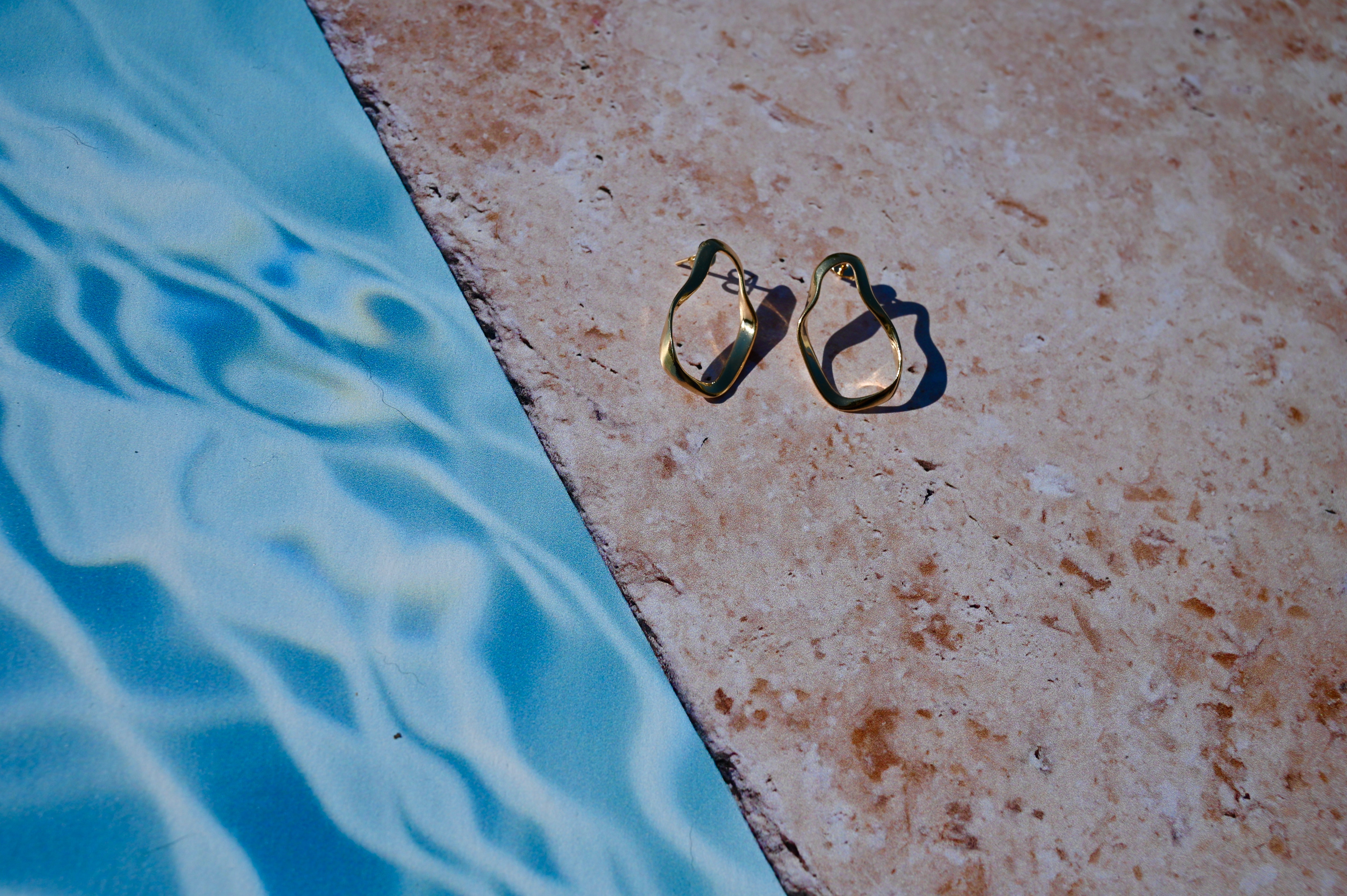 Asymmetric gold hoop earrings on a stone ledge by a pool