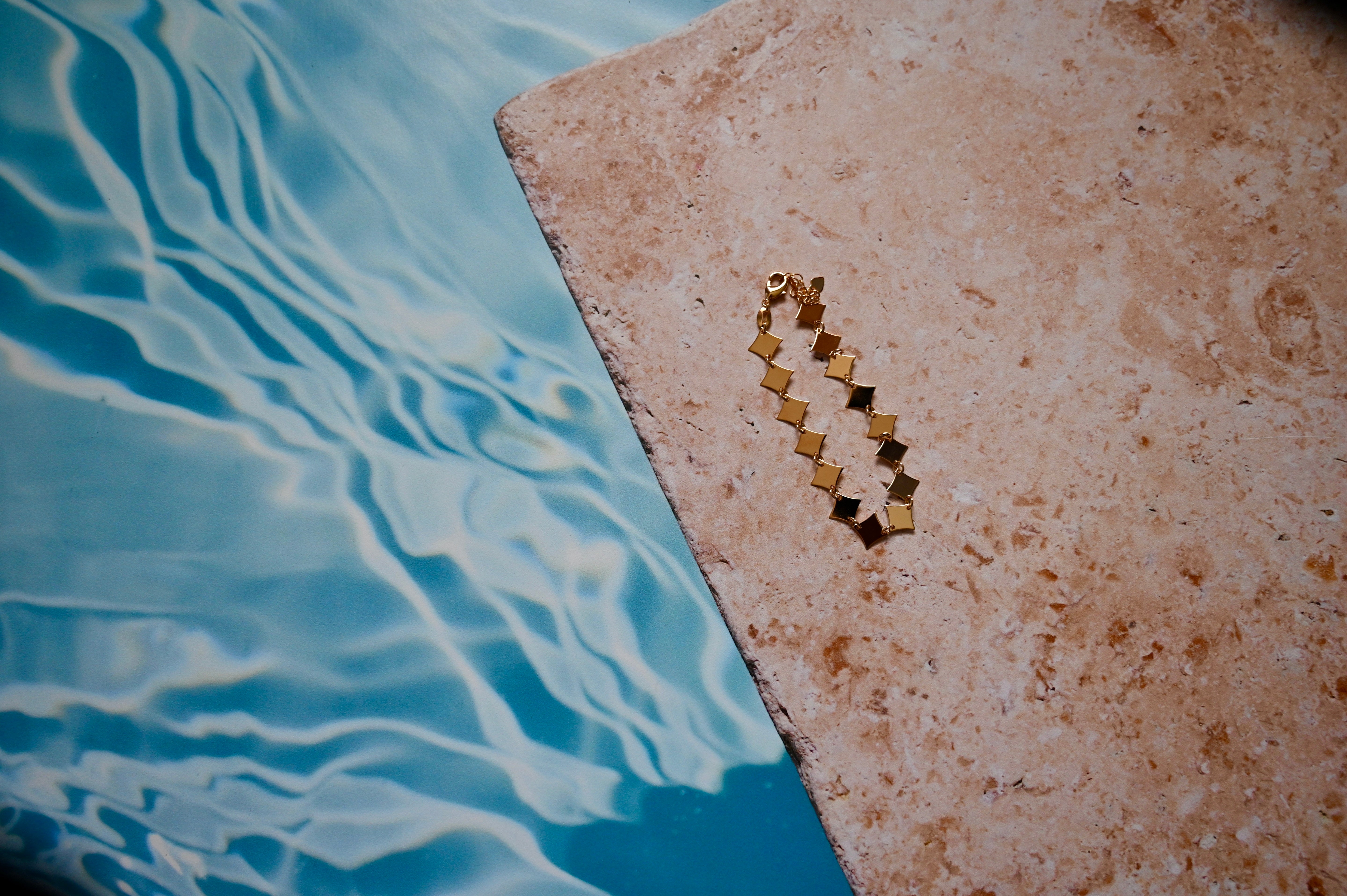 Gold bracelet with continuous diamond shapes on a stone ledge next to a pool